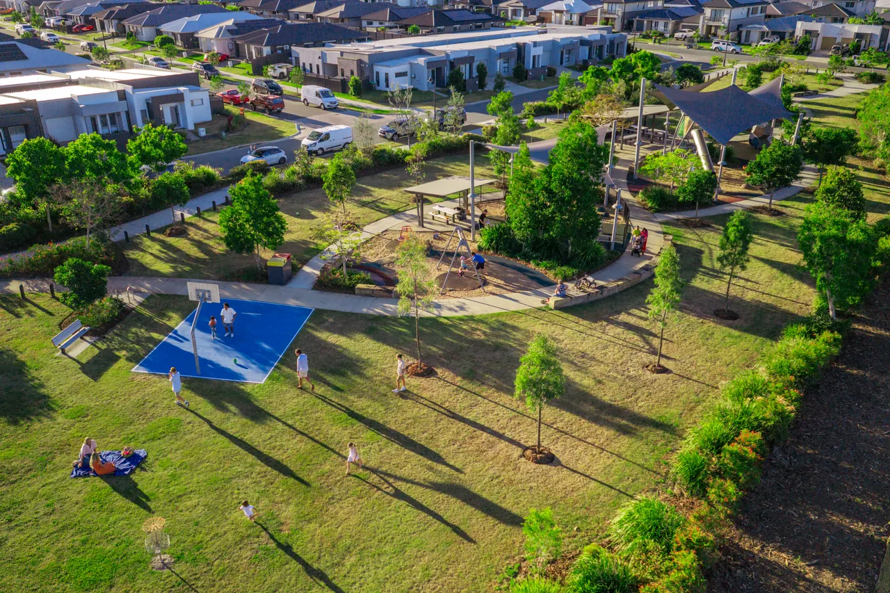 An aerial view of a neighbourhood park with a basketball court, playground and picnic shelter beside homes