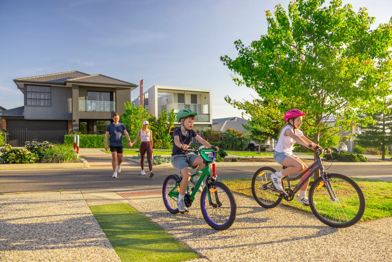Two children riding bikes along a footpath past new homes, with their parents walking behind